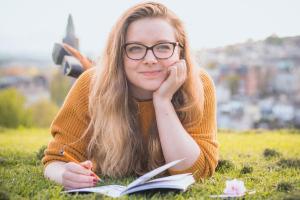 Eine versonnen lächelnde junge Frau liegt auf einer Wiese, vor sich ein Notizheft, in der Hand einen Stift. 