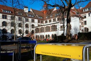 Innenhot der Alten PH unter blauem Himmel; im Vordergrund ein leuchtend gelber Stehtisch mit der Aufschrift PH HEIDELBERG.