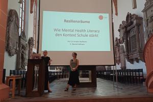 Prof. Dr. Annette Haußmann bei ihrem Vortrag in der Akademischen Mittagspause am Pult der Peterskirche. Neben ihr Rieke Deborah Koßmann. Hinter ihr eine Folie mit dem Titel ihres Vortrags.