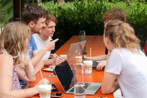 Eine gemischte Gruppe Studierender im Gespräch an einer Bierbank, vor sich Kaffee und Laptops.