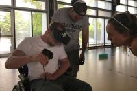 In einer Turnhalle treibt ein Student im Rollstuhl Sport mit VR-Brille und Controllern, 2 Studierende unterstützen