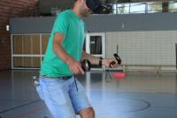 In einer Turnhalle balanciert ein Student mit VR-Brille und Controllern über eine umgedrehte Bank