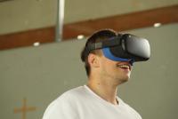 Lachender Student in einer Turnhalle beim Ausprobieren einer digitalen Sporteinheit mit VR-Brille