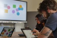 In einem Seminarraum arbeiten ein junger Mann und eine junge Frau an iPads, im Hintergrund ist ein Smartboard mit bunter Grafik erkennbar.