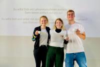 Drei lachende Lehramtsstudierende in HSE-Shirts vor dem Willkommensscreen des Lehramts-Cafés, in der Hand eine Tasse.