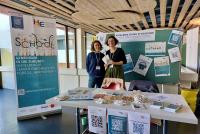 Margherita Maulella und Dr. Anuschka Holste-Massoth am mit Flyern und Merchandise ausgelegten HSE-Infostand in der Triplex-Mensa, im Hintergrund Plakate und Roll-ups.