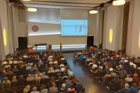 Dr. Christiane Wienand, Geschäftsführerin der HSE seitens der Universität und Leiterin des HSE-Prüfungsamts, auf der Bühne der Neuen Aula. Im Vordergrund mit Gästen voll besetzte Stuhlreihen.