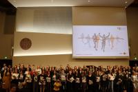Die Absolvent:innen der Studiengänge Master of Education Profillinie Lehramt Gymnasium sowie Master of Education Gerontologie, Gesundheit und Care auf der Bühne der Neuen Aula der Universität Heidelberg. In den Händen halten sie eine HSE-Mappe. Auf die Leinwand hinter ihnen ist eine stilisierte Grafik projiziert, die Läufer:innen beim Zieleinlauf zeigt.