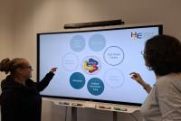 Die HSE-Mitarbeiterinnen Johanna Ruge und Dr. Anuschka Holste Massoth stehen vor einem eingeschalteten Smartboard und zeigen auf relevante Inhalte. Auf dem Smartboard sind in Kreisen die Begriffe "Lehre in den Verschränkungsmodulen", "Workshop", "toolbox Verschränkung" sowie "Fach-Sheet Fächer" zu lesen.