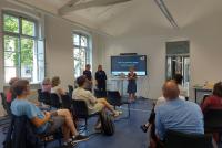 Teilnehmende des HSE-Terrassengesprächs sitzen wegen unbeständigen Wetters im HSE-Konferenzraum und schauen nach vorne auf ein Smartboard. Vor dem Smartboard stehen die drei Referent:innen Prof. Dr. Barbara Mittler, Dr. Odila Schröder und Dr. Daniel Schumacher.