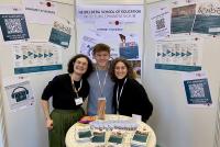Dr. Anuschka Holste-Massoth, Mathis Härtling und Margherita lachen in die Kamera. Sie stehen vor einer Stellwand, die voller Plakate mit Veranstaltungsankündigungen und Studieninformationen hängt. Vor ihnen steht ein Stehtisch, auf dem allerlei Flyer der HSE ausliegen.