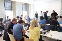 Einige Teilnehmende des Heidelberg Informatiklehrkräftetags sitzen in einem Seminarraum in kleinen Gruppen zusammen, haben aufgeklappte Laptops vor sich stehen und sind in Gespräche vertieft. Andere Teilnehmende stehen in kleineren Gruppen zusammen und sprechen miteinander.