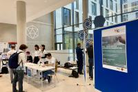 Im Foyer des Mathematikons stehen Tische für das Tagungsbüro des Heidelberger Informatiklehrkräftetags, an denen Mitglieder des HSE-Teams die Unterlagen für die neu ankommenden Teilnehmenden ausgebreitet haben. Eine Teilnehmerin nähert sich gerade den Tischen und dem Personal des Tagungsbüros. Rechts im Vordergrund sieht man eine Stellwand, auf der das Plakat des HILT hängt.