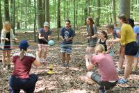Studierende stehen im Kreis auf einer Lichtung im Wald. Alle von ihnen halten jeweils Enden von zwei langen Schnüren in den Händen; die jeweils anderen Enden der Schnüre sind alle zusammen verknotet. Dieser Knoten befindet sich in der Mitte des Personenkreises und auf ihm liegt eine kleine Erdkugel. Die Studierenden versuchen, das Konstrukt der Schnüre so auszubalancieren, dass die Erdkugel nicht herunterfällt.