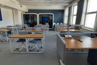 Das Foto zeigt ein modernes finnisches Klassenzimmer mit Teppichboden. Es ist ausgestattet mit vielen Einzeltischen sowie rollfähigen, bequemen Schreibtischstühlen, womit sich die Sitzordnung der Schüler:innen leicht verändern lässt. Im Hintergrund sind zwei geräuschdämpfende Arbeitskabinen zu erkennen, in die sich die Schüler:innen für konzentriertes Arbeiten zurückziehen können. Auf der linken Seite des Zimmers hängt ein Smartboard an der Wand.