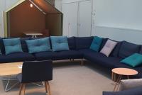 Das Foto zeigt einen großen Raum mit ausladender blauer Sitzgruppe in L-Form und Tischen. Dahinter eine Art offenes Häuschen mit Tisch, welches für Meetings oder konzentriertes Arbeiten individuell genutzt werden kann. Es handelt sich dabei um ein finnisches Lehrerzimmer an einem großen Gymnasium, das von über 1000 Schüler:innen besucht wird. Die Architektur des Raumes ist luftig und offen. Die verwendeten Materialien, wie Teppichboden, Kissen und Stoffe sorgen für eine geräuschmindernde Umgebung.