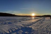 Das Foto zeigt einen klaren, blauen Himmel und eine weite Schneefläche, die durch die untergehende Sonne in ein kühles, aber gleißendes Licht getaucht wird. In der verschneiten Fläche zeichnen sich schemenhaft eine Eisfläche und Skispuren ab. Menschen spazieren über dem zugefrorenen See in Jyväskylä, der im Winter als Sport- und Freizeitort genutzt wird.