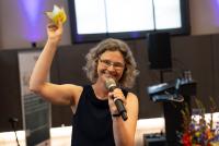 Dr. Christiane Wienand hält nach dem Science Contest die vier bunten Abstimmungskärtchen in die Höhe. In der anderen Hand hält sie ein Mikrofon.
