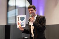 Dr. Sebastian Mahner am Mikrofon auf der Bühne der Festhalle, in der rechten Hand ein Schild mit der Aufschrift "I love HSE"