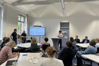 Teilnehmende des Zukunftsforums sitzen an Tischen gruppiert im Konferenzraum der HSE. Am Smartboard steht Dr. Daniel Vetter, links daneben Dr. Dennis Dietz und Dr. Lina Pranaityte. Der Screen zeigt "Exemplarische studentische Perspektiven" zu zukunftsgerichteten Leitfragen.