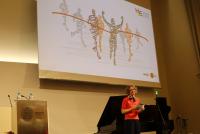 Dr. Christiane Wienand auf der Bühne der Neuen Universität. Im Hintergrund eine Grafik, die stilisierte Menschen beim Zieleinlauf zeigt.