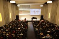 Prof. Dr. Michael Haus am Flügel auf der Bühne der Neuen Aula. Im Vordergrund die voll besetzten Stuhlreihen.