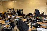 Blick in ein Klassenzimmer mit Tischen, Stühlen und auf diesen verteilten Rucksäcken.