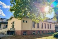 Das HSE-Gebäude in der Voßstraße 2 unter blauem Himmel, rechts davor eine grüne Wiese mit großem Baum, durch den die Sonne bricht.
